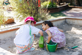 Two children playing in a sandbox, one in a pink hat with a shovel, the other in a patterned dress with a green bucket, surrounded by greenery.