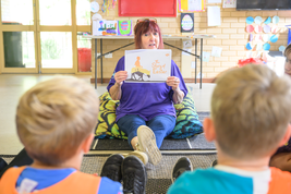 A woman sits on a pillow, showing a book titled "The Story of Easter" to children in a classroom setting.
