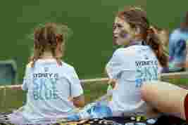Two girls with braided hair sit on grass wearing "Sydney is Sky Blue" T-shirts, watching an event with a face painting on one girl.