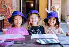 Three young girls wearing purple hats, smiling at the camera, seated at a table with art supplies in an outdoor setting.