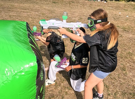 Three children in black vests and protective gear participate in a gel blaster game, taking cover behind a green inflatable barrier.