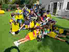 A group of children wearing goggles and yellow or blue vests hold toy guns while posing on a lawn in a playful, outdoor setting.