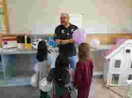 A teacher with purple balloons engages with three young girls in a classroom near a dollhouse.