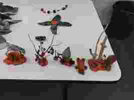 Natural craft made of leaves, twigs, pinecones, and orange clay on a white table, with a trash bin in the background.