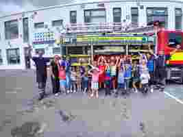 A group of excited children and adults standing in front of an open fire truck, with a building in the background on a cloudy day.