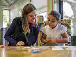 Smiling woman and girl sitting at a table, playing a card game in a brightly lit room with bookshelves in the background.