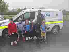 A group of young children stands smiling in front of a Garda vehicle with a Garda officer on an overcast day.