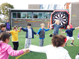 Children holding hands in a circle on a school playground, playing near a giant dartboard.