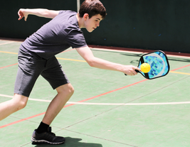 A person in athletic attire plays pickleball on an indoor court, reaching out with a paddle to hit a yellow ball.
