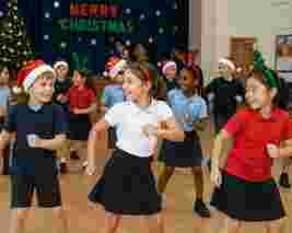 Children in festive attire, wearing Santa hats and reindeer antlers, dance happily in a school hall decorated for Christmas.