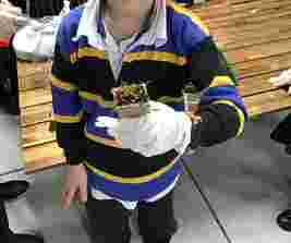 Child in blue and yellow striped sweater holding a small pot with sprouts, wearing gloves, standing by a wooden table.