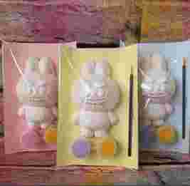 Three packaged Labu(bu) bunny-shaped ceramic figures with paintbrushes and paint pots, set against a colourful wooden background.