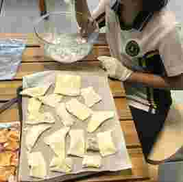 A person preparing pastry, shaping dough pieces on parchment paper, with a bowl of filling mixture on a wooden table.