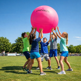 Children playing with a large pink ball outdoors on a sunny day, surrounded by green grass and trees, wearing casual athletic clothing.