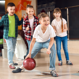 Four children at a bowling alley, smiling as one boy in a white shirt prepares to roll a red bowling ball down the lane.