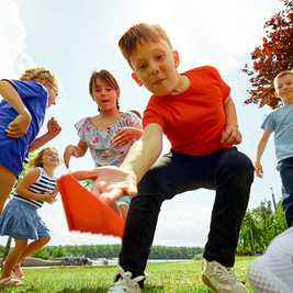 Children play together outdoors on grass, focused on a boy in a red shirt reaching out with an orange cone. Sunny day, trees in the background.