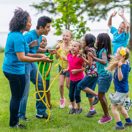 Children and adults joyfully engage in a group activity involving a rope on a grassy field, with everyone smiling and cheering.