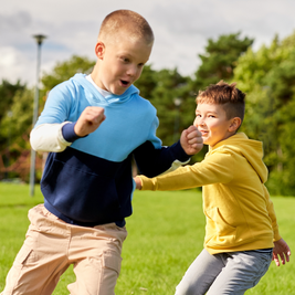 Two boys wearing colorful hoodies play and run joyfully on a grassy field, with trees and a lamp post in the background.