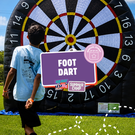 Boy playing foot dart at a summer camp event, with a giant inflatable dartboard in the background.