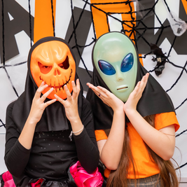 Two people wearing Halloween masks—one a smiling pumpkin and the other an alien—pose in front of a spooky, web-decorated backdrop.