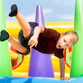 A child in a red shirt delightfully jumps on a colorful inflatable play structure, surrounded by bright green, purple, and yellow cones.