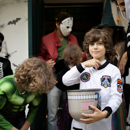 Children in Halloween costumes, including a space suit and superhero, gather at a decorated door with a masked adult in the background.