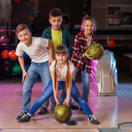 Four children at a bowling alley, smiling and posing with bowling balls on the lane.