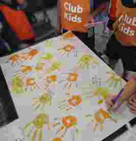Children wearing "Club Kids" vests paint handprints on paper, writing names and messages in colorful inks.