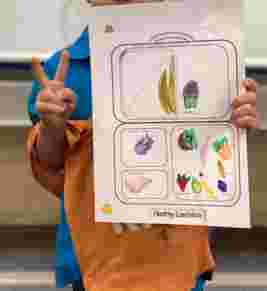 Child holding a "Healthy Lunchbox" drawing, featuring sketches of a banana, cupcake, fruits, and vegetables, making a peace sign.