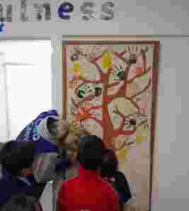A group of children observe a large paper tree mural with colorful handprints in a classroom under the word "wellness."