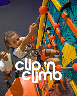 Child climbing a colorful indoor wall with orange holds at Clip 'n Climb, supervised by an adult below.