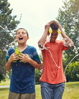 Two boys play with water balloons, laughing as water splashes over them on a sunny day in a grassy park.