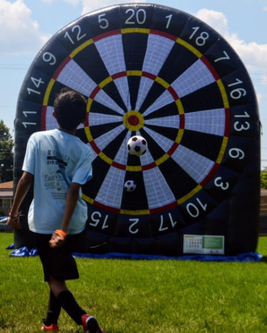 A boy in a light blue shirt kicks a soccer ball towards a large inflatable dartboard on a grassy field.