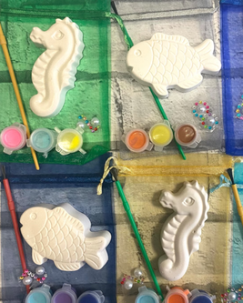 Unpainted seahorse and fish plaster figures with paintbrushes, colorful paints, and small beads on colorful mesh bags.