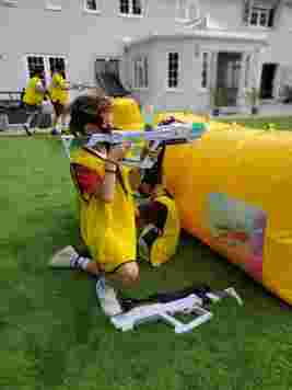 Children wearing yellow vests engage in a laser tag game on a grassy area with inflatable barriers and toy guns.