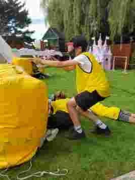 Kids wearing yellow vests and goggles play with toy guns behind inflatable barriers on a grassy field.