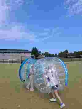 Two people in inflatable bumper balls on a grassy field, with a building and trees in the background under a clear blue sky.