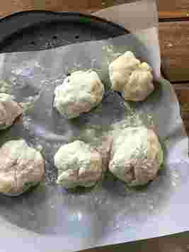 Six dough balls resting on parchment paper on a baking tray, dusted with flour, ready for baking.