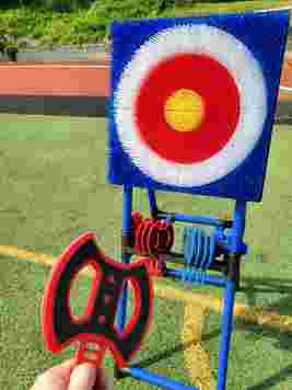 Hand holding a red and black throwing axe aimed at a colorful target on a grassy field.