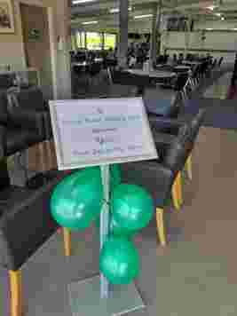Sign reading "North Beach Bowling Club Welcomes Green Day on the Green" with green balloons, inside a room with chairs and lights.