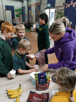 A woman pours a smoothie for a group of children in a classroom. The table holds bananas and bags of mixed berries.