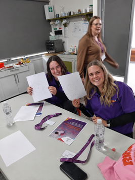 Two smiling women in purple shirts hold up drawings at a table with books and water bottles; another person stands in the background.