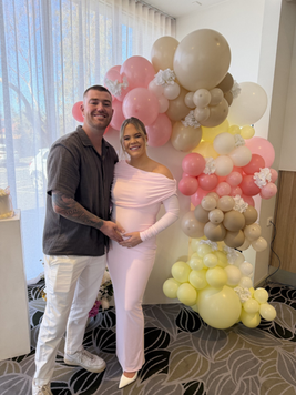A smiling couple poses in front of a colorful balloon arch; the woman is wearing a pink dress, and they are holding her baby bump.