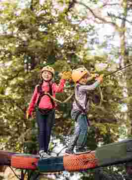Two children in safety gear, including helmets and harnesses, navigate a high rope course outdoors, surrounded by trees.