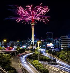 A view of a busy highway with lights streaking down from the fast cars and a lit up city in the background with a tall pointy structure in the middle that is shooting pink fireworks from the top of the structure into the night sky