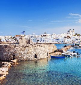 A calm harbor with clear blue water and a stone wall with many densely packed white homes in the background on a clear blue day