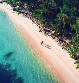 An aerial view of two people walking along the shore of a beach between crystal blue water and a lush green forest