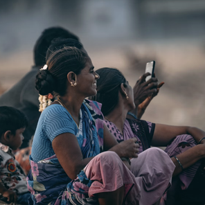 A group of women and a child sit outdoors, all are relaxed. One woman is holding a smartphone. The scene is informal and candid.