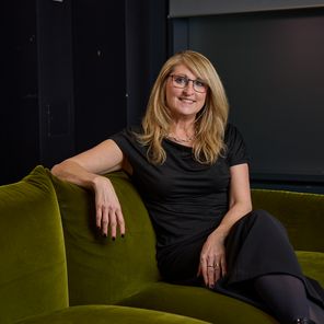Anne Stagg, CEO of Digitas UK with long blonde hair and glasses sits relaxed on a green sofa, wearing a black outfit, with bookshelves in the background.