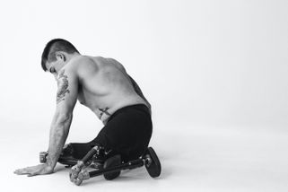 Ein Mann ohne Hemd, mit Tätowierungen und Beinprothesen sitzt mit abgewandtem Gesicht in einer minimalistischen Umgebung auf dem Boden.